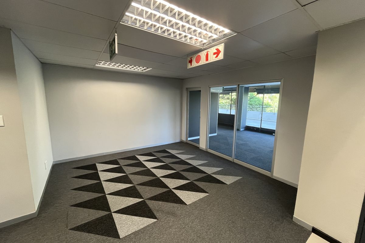 A bright, empty office space featuring modern carpet tiles, recessed fluorescent lighting, and a sliding glass door leading to an outdoor area.