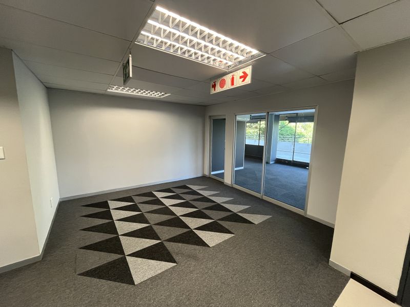 A bright, empty office space featuring modern carpet tiles, recessed fluorescent lighting, and a sliding glass door leading to an outdoor area.