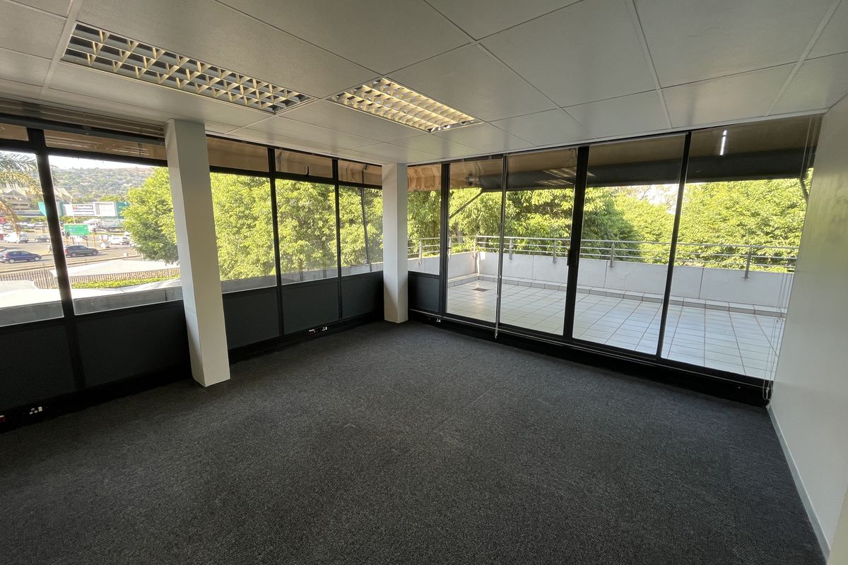 A bright, empty office space featuring large floor-to-ceiling windows, carpeted flooring, and direct access to a private balcony with exterior views.