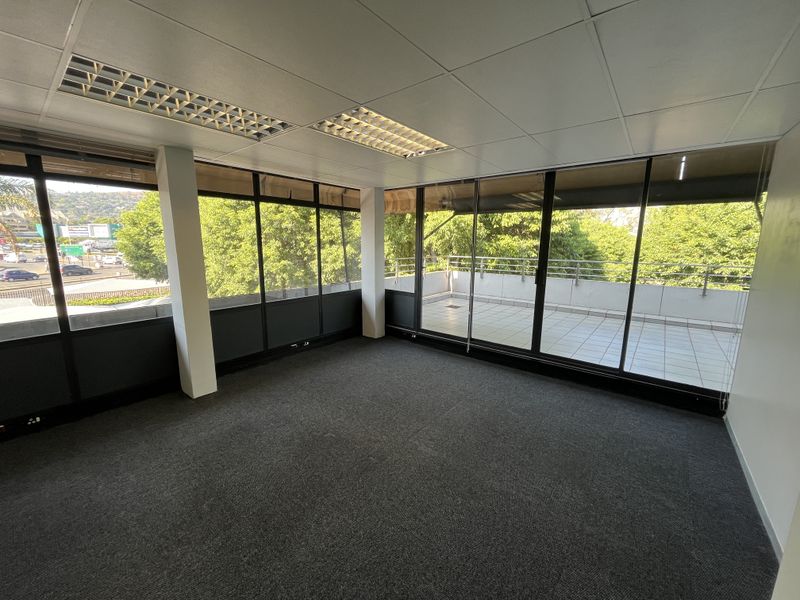 A bright, empty office space featuring large floor-to-ceiling windows, carpeted flooring, and direct access to a private balcony with exterior views.