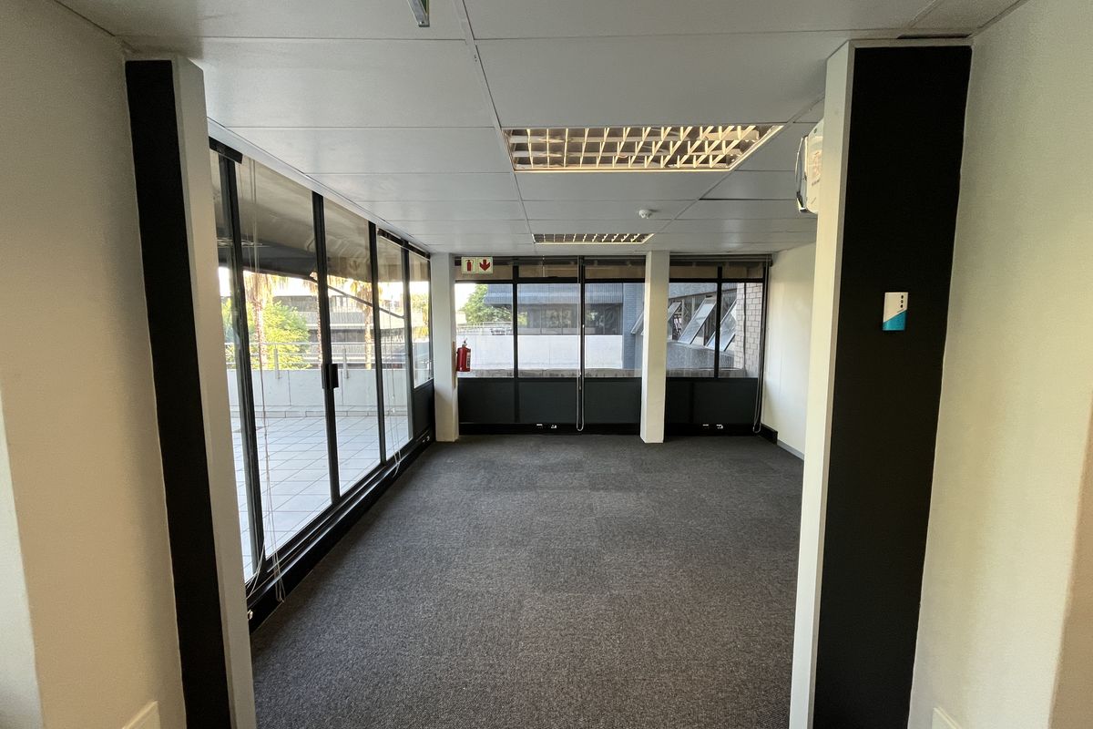 An empty, carpeted office space featuring a suspended ceiling, recessed lighting, and large windows providing natural light and access to an outdoor balcony area.