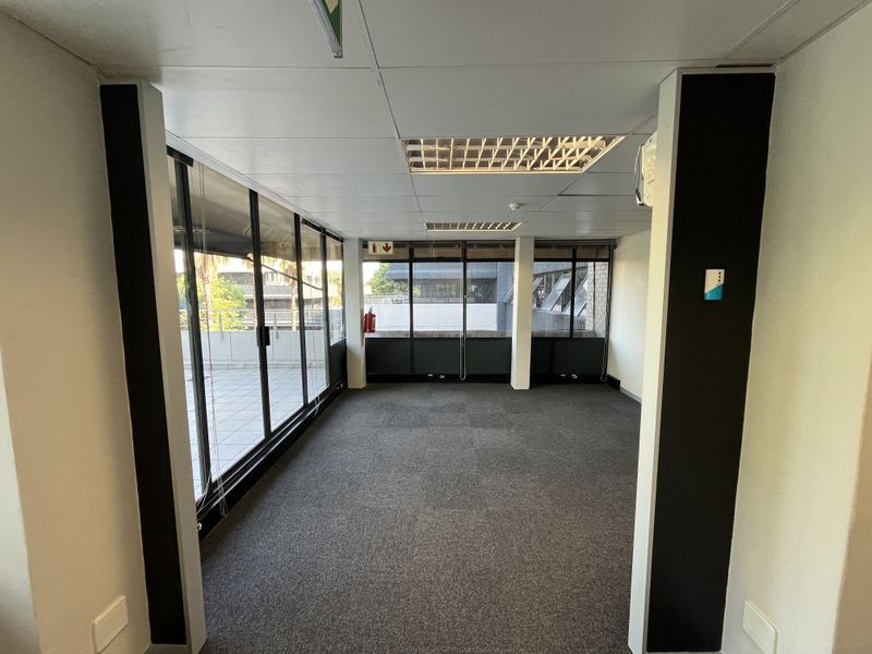 An empty, carpeted office space featuring a suspended ceiling, recessed lighting, and large windows providing natural light and access to an outdoor balcony area.