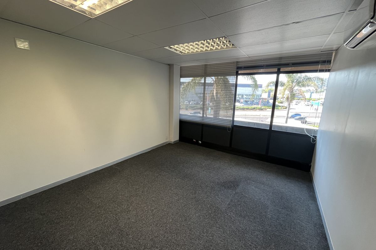 A clean, empty office space featuring carpeted flooring, a suspended ceiling with recessed lighting, and a wall-mounted air conditioning unit with large windows.