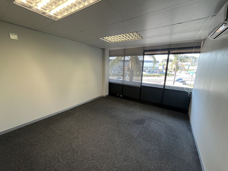 A clean, empty office space featuring carpeted flooring, a suspended ceiling with recessed lighting, and a wall-mounted air conditioning unit with large windows.