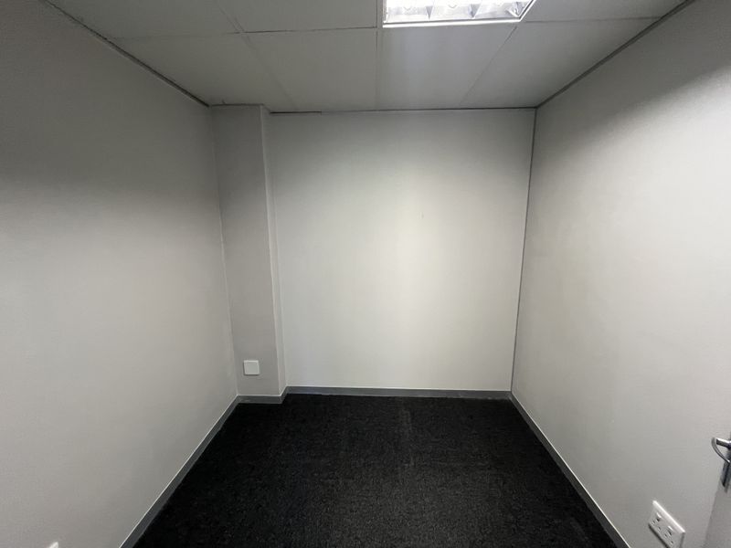 A vacant, compact office space featuring neutral walls, dark carpeting, and a suspended ceiling with integrated lighting.