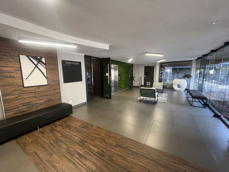 Modern office building lobby featuring tiled flooring, a wood-look feature wall, elevator access, and a glass curtain wall providing natural light.