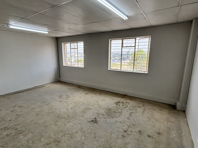 An empty industrial office space featuring a suspended ceiling, fluorescent lighting, and security-barred windows, with concrete flooring in need of refurbishment.
