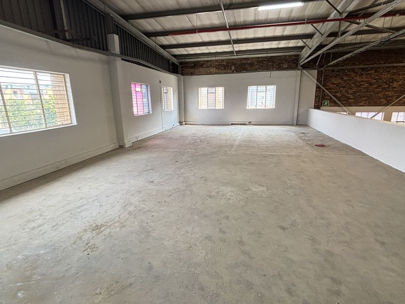 Spacious industrial mezzanine area featuring exposed steel trusses, concrete flooring, and natural light through security-barred windows, suitable for office or storage use.