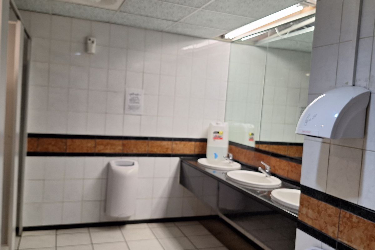 A functional office restroom featuring multiple washbasins, large vanity mirrors, tiled surfaces, and a drop ceiling with integrated lighting.