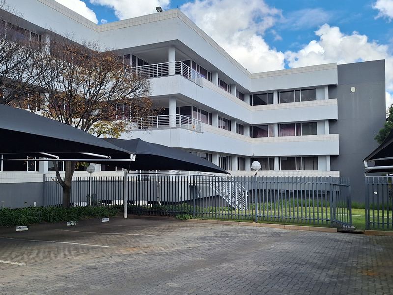 Exterior view of a multi-story office building featuring a paved parking area with shade cloth carports and secure perimeter fencing.