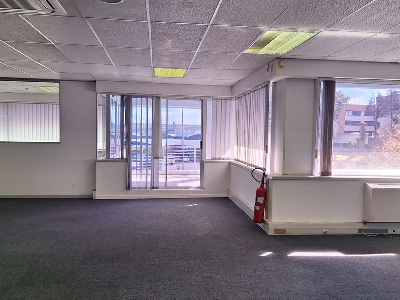 A spacious, empty office area featuring carpeted floors, a suspended ceiling with recessed lighting, large windows, and access to an outdoor balcony.