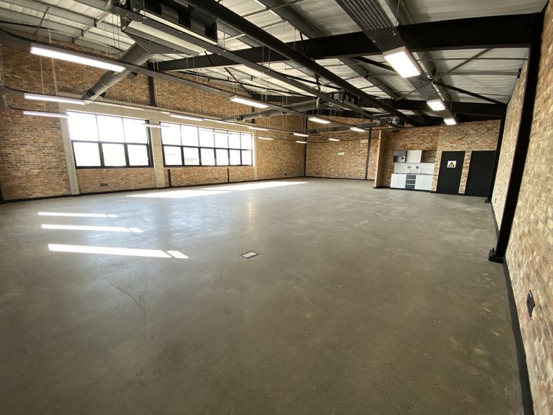 Spacious open-plan industrial unit featuring exposed brick walls, polished concrete flooring, high ceilings with exposed steel trusses, and large windows for natural light.