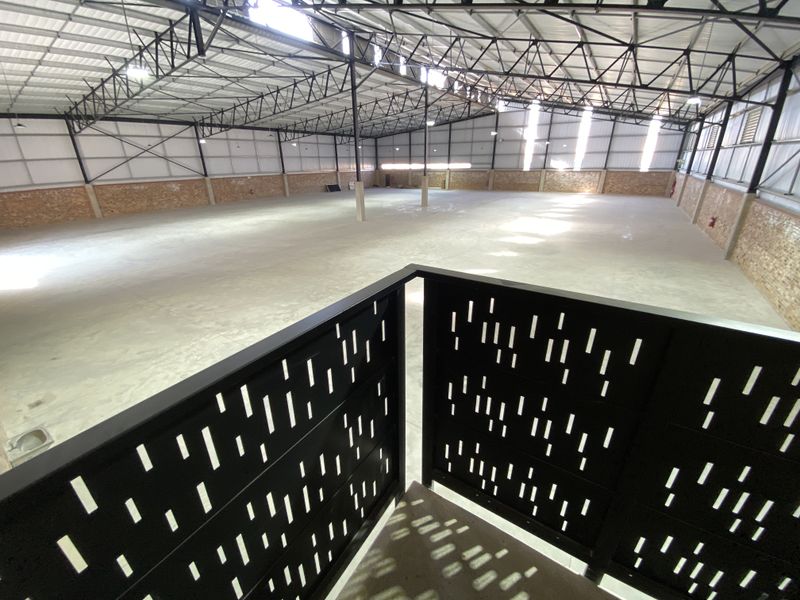 A spacious industrial warehouse interior featuring high ceilings with exposed steel trusses, concrete flooring, and a decorative metal mezzanine balustrade.
