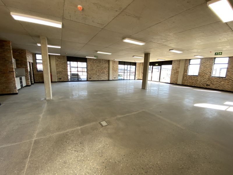 Spacious open-plan industrial office space featuring exposed brick walls, polished concrete flooring, and large windows providing ample natural light.
