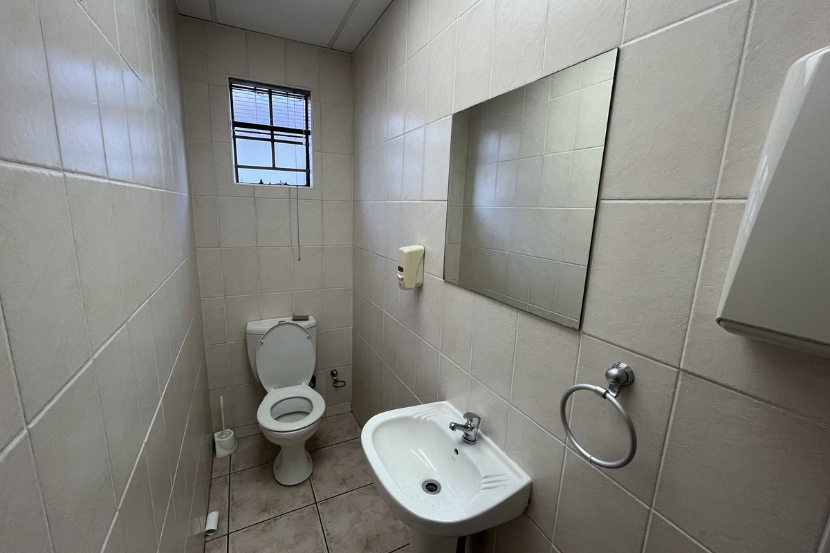A functional office restroom featuring full-height wall tiling, a pedestal basin, a wall-mounted mirror, and a standard toilet suite.