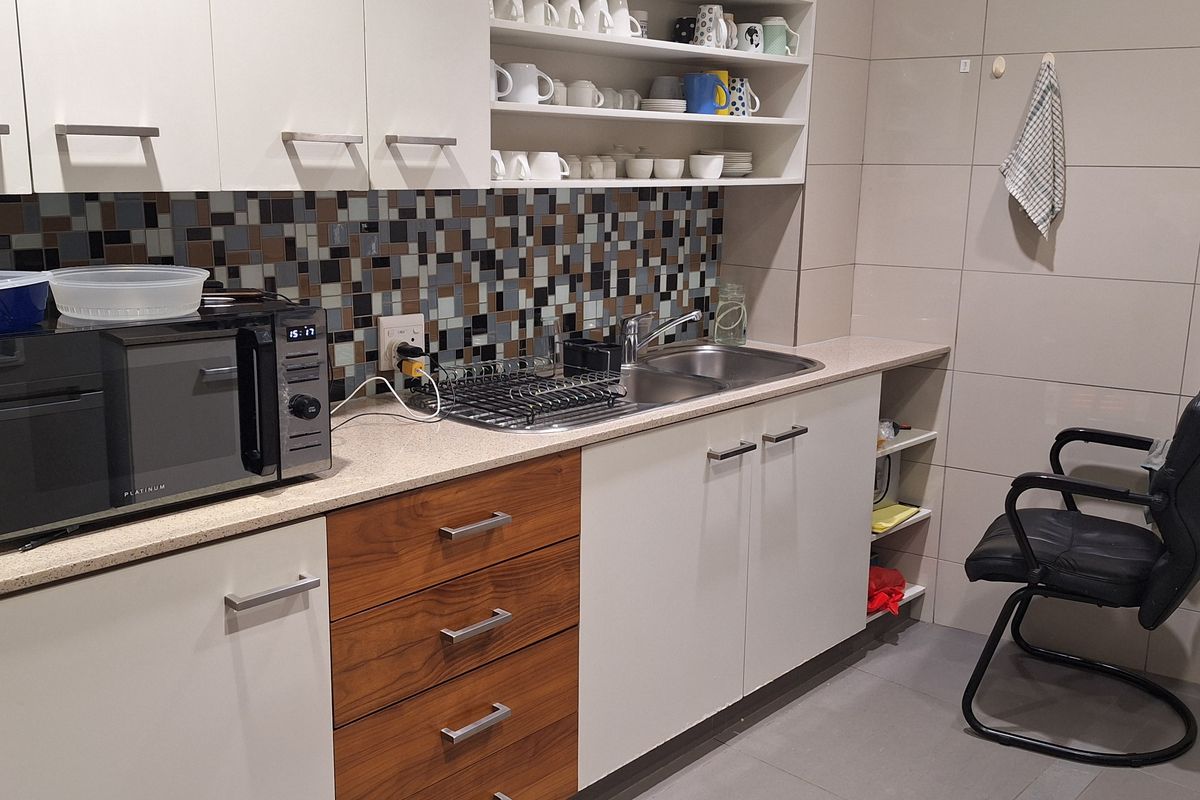 A functional office kitchenette featuring modern white and wood-grain cabinetry, a mosaic tile backsplash, stainless steel sink, and tiled flooring.