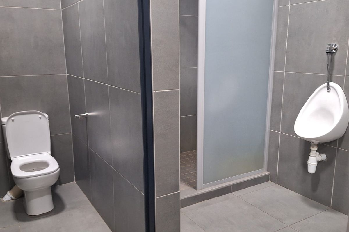 A modern commercial bathroom featuring grey tiled walls and floors, equipped with a toilet, a wall-mounted urinal, and a glass-enclosed shower stall.