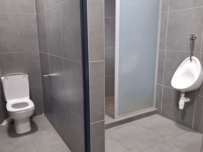 A modern commercial bathroom featuring grey tiled walls and floors, equipped with a toilet, a wall-mounted urinal, and a glass-enclosed shower stall.