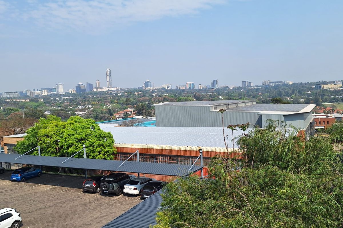 Aerial view of a commercial property featuring a paved parking area with covered carports, metal roofing, and solar panels overlooking the city skyline.