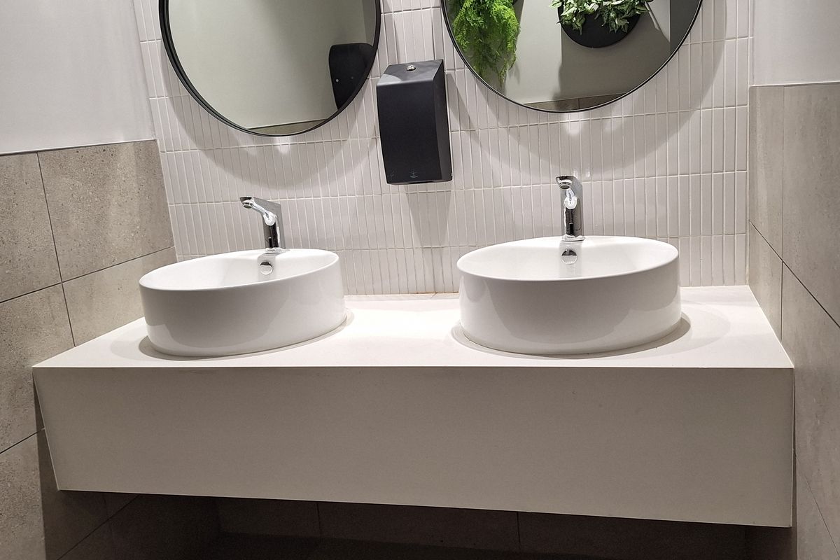 Modern office bathroom featuring a floating vanity with dual vessel sinks, contemporary wall-mounted faucets, and clean tiled finishes.