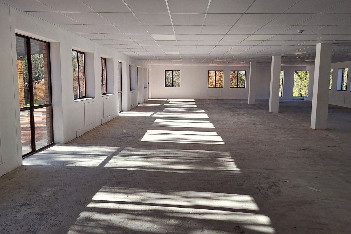A large, open-plan office space featuring a suspended ceiling, structural columns, and abundant natural light through multiple windows and a glass sliding door.