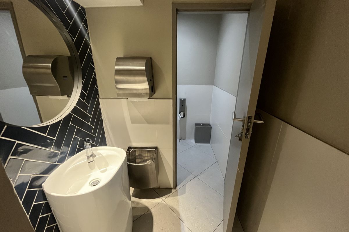 Modern office restroom featuring a pedestal sink, herringbone-patterned tiled backsplash, circular mirror, and stainless steel wall-mounted hand dryer.