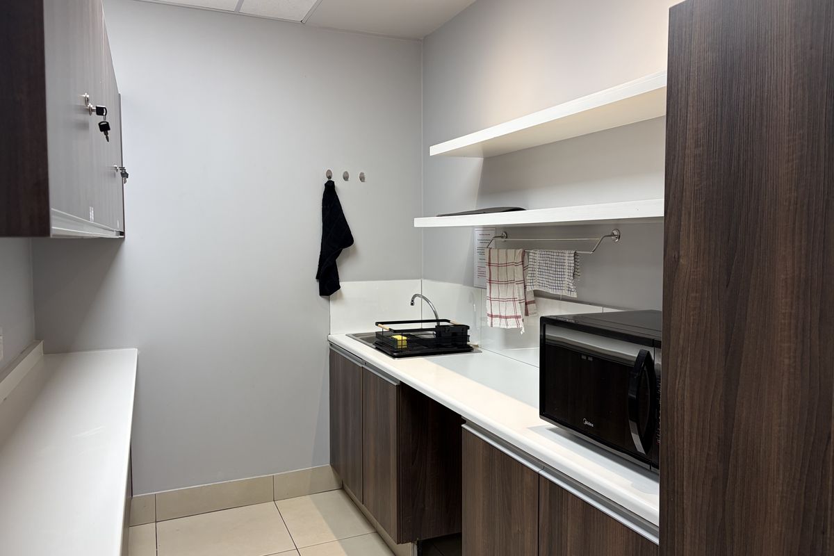 A compact commercial kitchenette featuring dark wood cabinetry, white countertops, open shelving, and a built-in microwave.