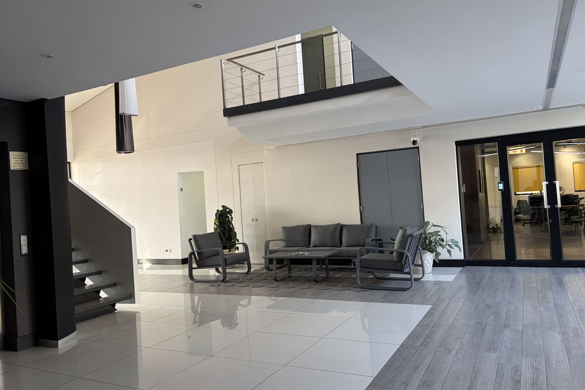 Spacious commercial lobby featuring a double-volume ceiling, mezzanine level with metal railing, polished tile flooring, and glass-paneled entrance doors.