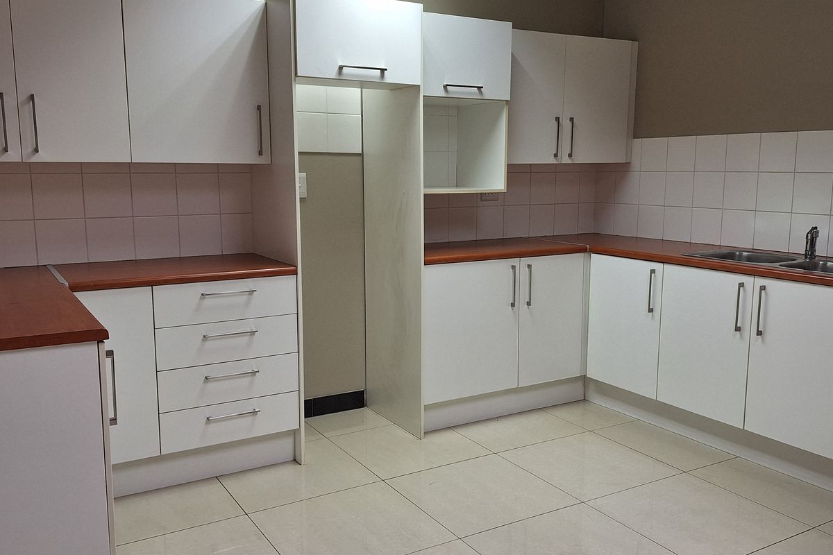 A clean office kitchenette featuring white cabinetry, wood-look laminate countertops, tiled splashbacks, and a stainless steel sink.