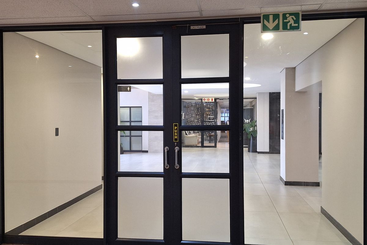 A professional office entrance featuring modern glass double doors with black frames, leading into a bright, tiled lobby area with recessed ceiling lighting.