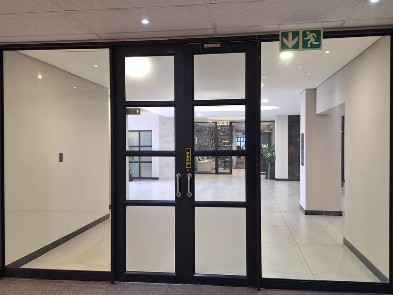 A professional office entrance featuring modern glass double doors with black frames, leading into a bright, tiled lobby area with recessed ceiling lighting.