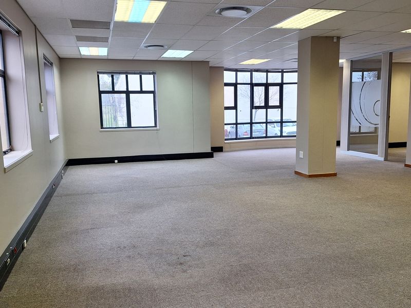 A spacious, open-plan office area featuring large windows, a central structural column, and a suspended ceiling with integrated lighting.