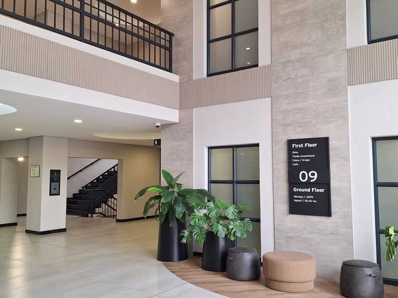 Modern double-volume office building lobby featuring tiled flooring, a mezzanine level with black metal railings, and integrated tenant directory signage.