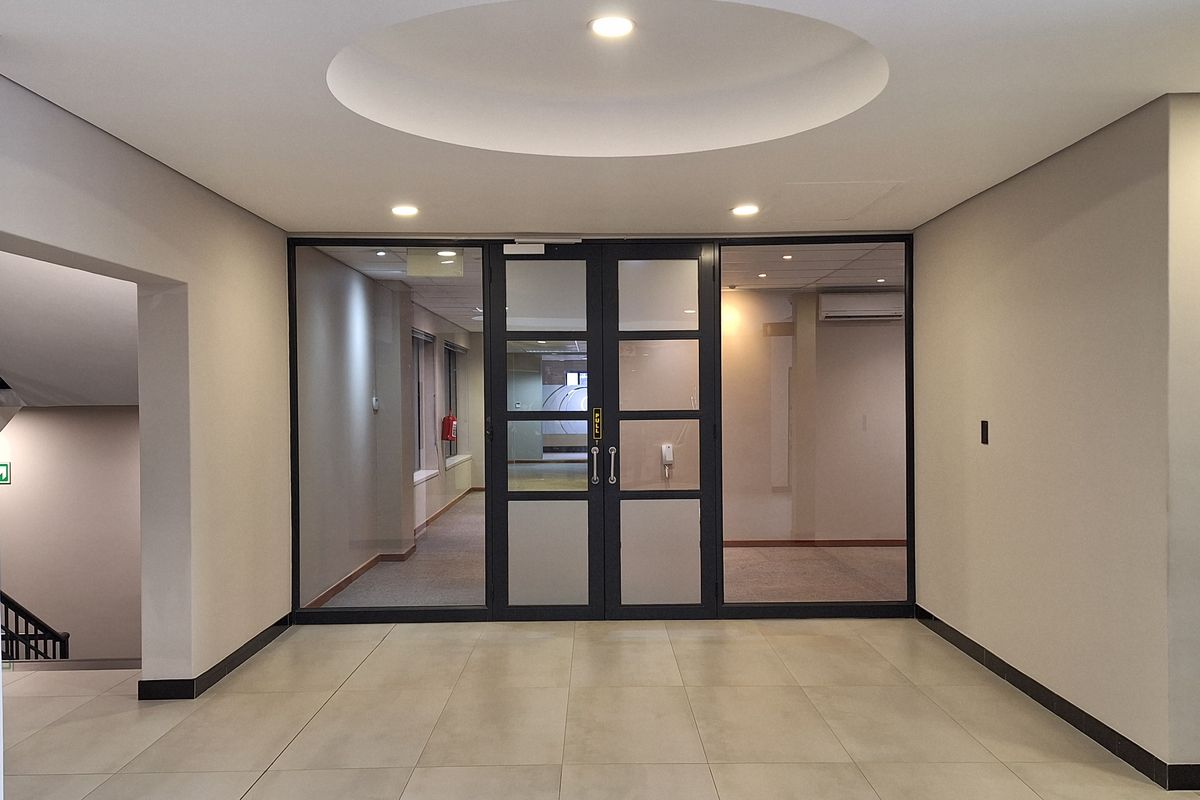 A bright office entrance lobby featuring tiled flooring, a recessed ceiling detail with integrated lighting, and glass-paneled double doors leading into the office space.