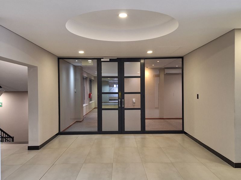 A bright office entrance lobby featuring tiled flooring, a recessed ceiling detail with integrated lighting, and glass-paneled double doors leading into the office space.