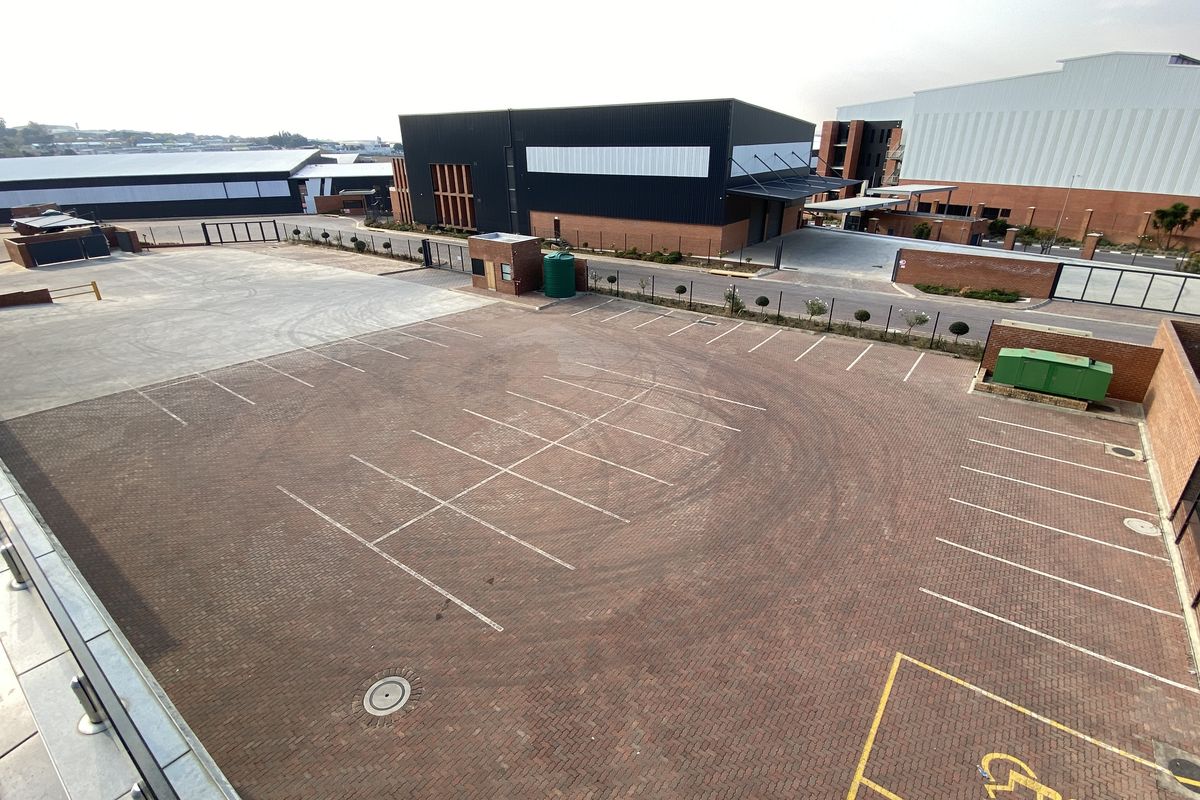 An elevated aerial view of a paved industrial parking lot and loading area, featuring marked parking bays and surrounding warehouse structures.