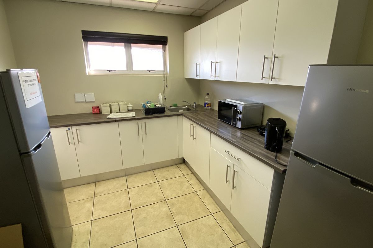 A functional kitchenette featuring white cabinetry, dark laminate countertops, a stainless steel sink, and tiled flooring in an industrial office setting.