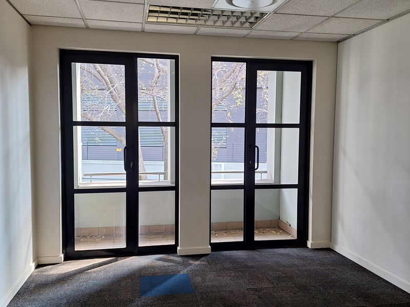 An empty office space featuring carpeted flooring, a suspended ceiling with integrated lighting, and dual glass doors leading to an outdoor area.