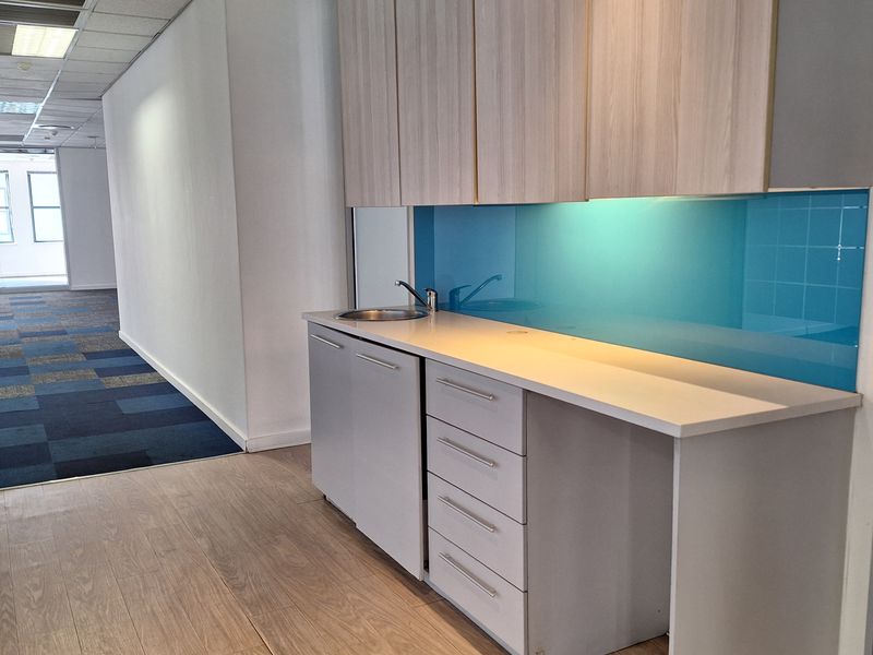 A modern office kitchenette featuring light wood-finish cabinetry, a blue glass splashback, and a compact sink, set within a clean, professional workspace.