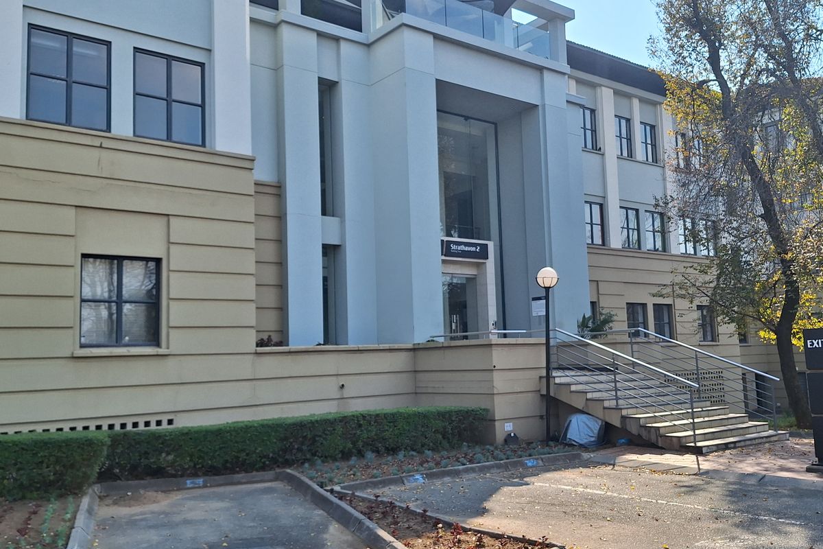 Front elevation of a modern office building featuring a glass entrance, balcony, and paved parking area with landscaped garden beds.