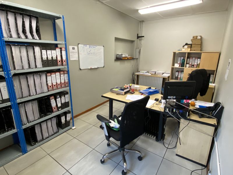 A functional industrial office space featuring tiled flooring, fluorescent lighting, and a wall-mounted whiteboard, suitable for administrative or operational tasks.