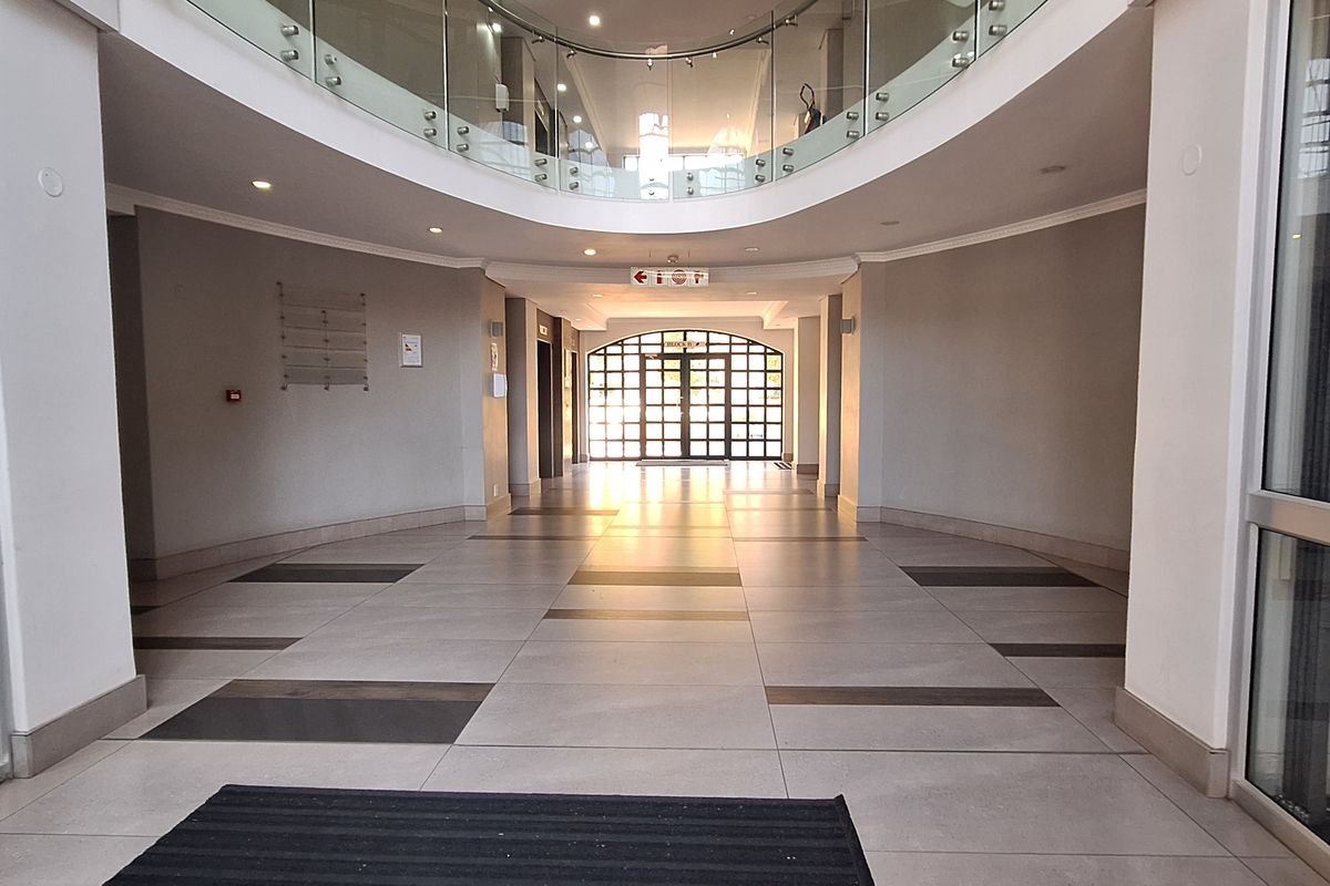 Spacious office building entrance lobby featuring tiled flooring, a mezzanine level with glass balustrade, and large glass entrance doors.