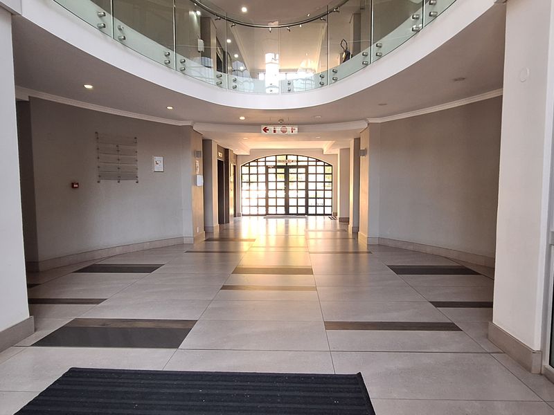 Spacious office building entrance lobby featuring tiled flooring, a mezzanine level with glass balustrade, and large glass entrance doors.