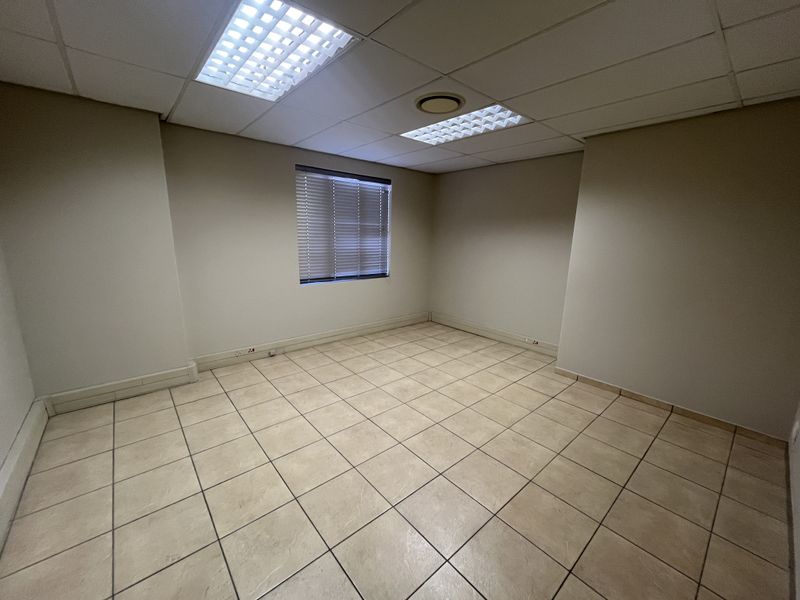 A clean, empty office space featuring ceramic floor tiles, a suspended ceiling with recessed lighting, and a window fitted with venetian blinds.