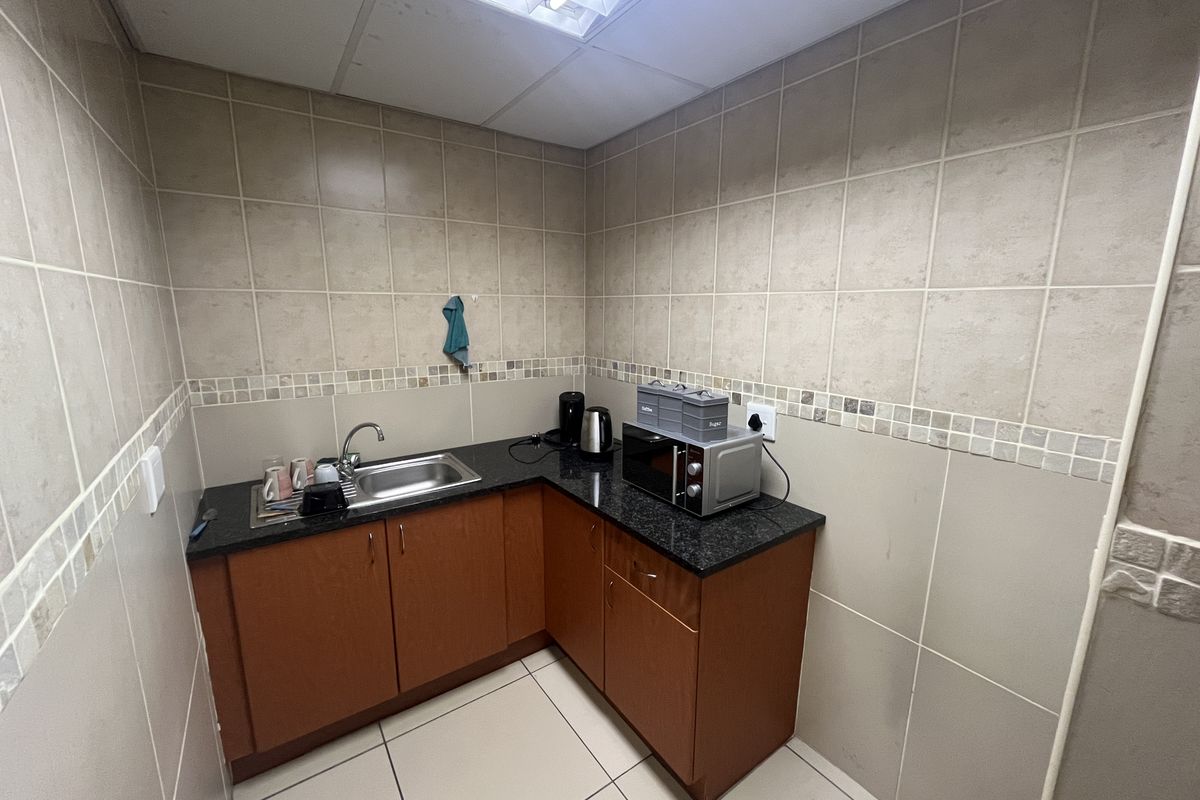 A compact office kitchenette featuring l-shaped cabinetry, granite countertops, a stainless steel sink, and full-height wall tiling.