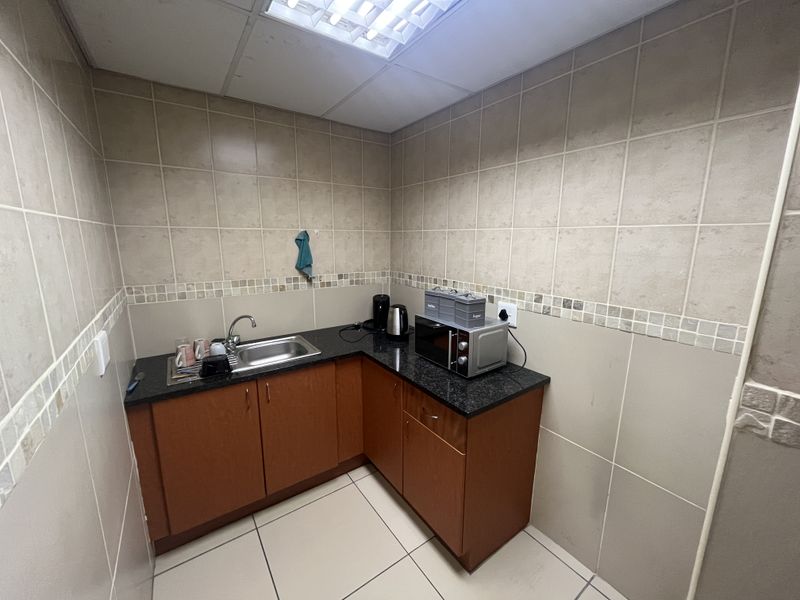 A compact office kitchenette featuring l-shaped cabinetry, granite countertops, a stainless steel sink, and full-height wall tiling.