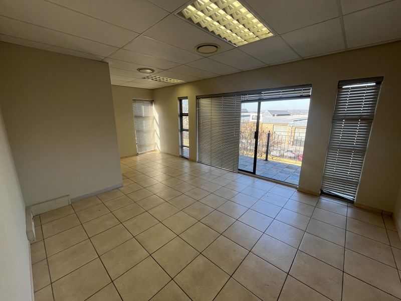 A bright, empty office space featuring ceramic tile flooring, suspended ceilings with recessed lighting, and a sliding glass door leading to a private balcony.