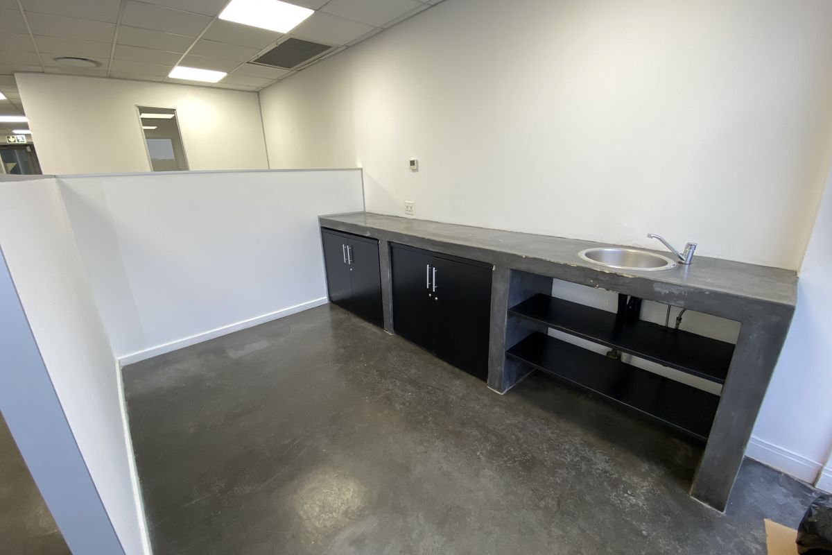 A functional office kitchenette featuring a concrete countertop, stainless steel sink, and black cabinetry, set within a clean, open-plan workspace.