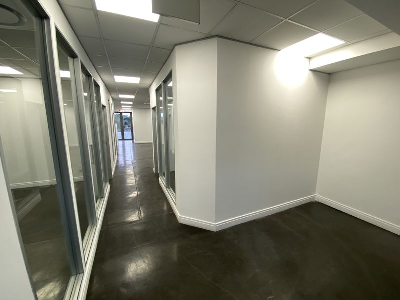 A modern, well-lit office hallway featuring polished concrete floors, suspended ceiling tiles, and glass partition walls leading to private office spaces.