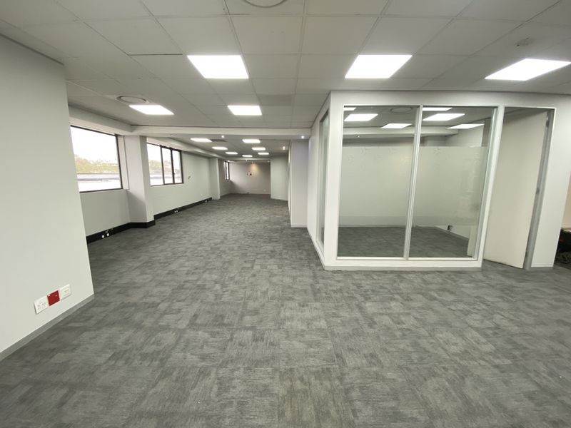 Spacious open-plan office area featuring carpet tiles, a suspended ceiling with recessed lighting, and a glass-partitioned meeting room.